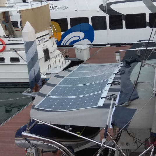 Flexible solar panels mounted on the canvas dodger or cover of a sailboat docked in a marina, with a ferry and another boat nearby.
