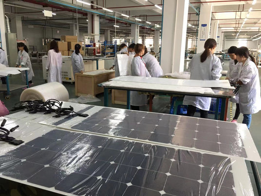 Workers in white lab coats assembling or inspecting flexible solar panels on tables in a clean factory environment.