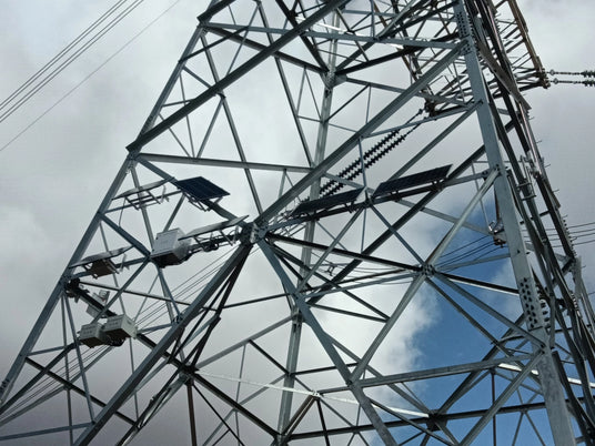 A power transmission tower has small solar panels and white junction boxes installed on its internal structure.