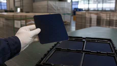 A gloved hand holds a square 125mm IBC back-contact solar cell above a tray of surplus stock in a warehouse.