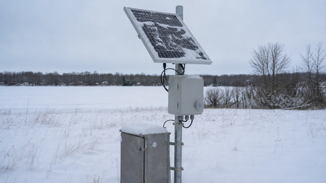 A solar-powered remote monitoring station in a snowy winter field, with a tilted panel partly covered in snow and a weatherproof enclosure on a pole.