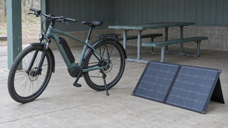 An unbranded electric bicycle and an unfolded portable solar panel stand at a dry rest stop, illustrating stationary charging planning without a claimed approved battery connection.