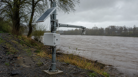 A solar-powered flood-monitoring station on a riverbank under an overcast stormy sky, with a radar water-level sensor arm extended over a swollen muddy river.