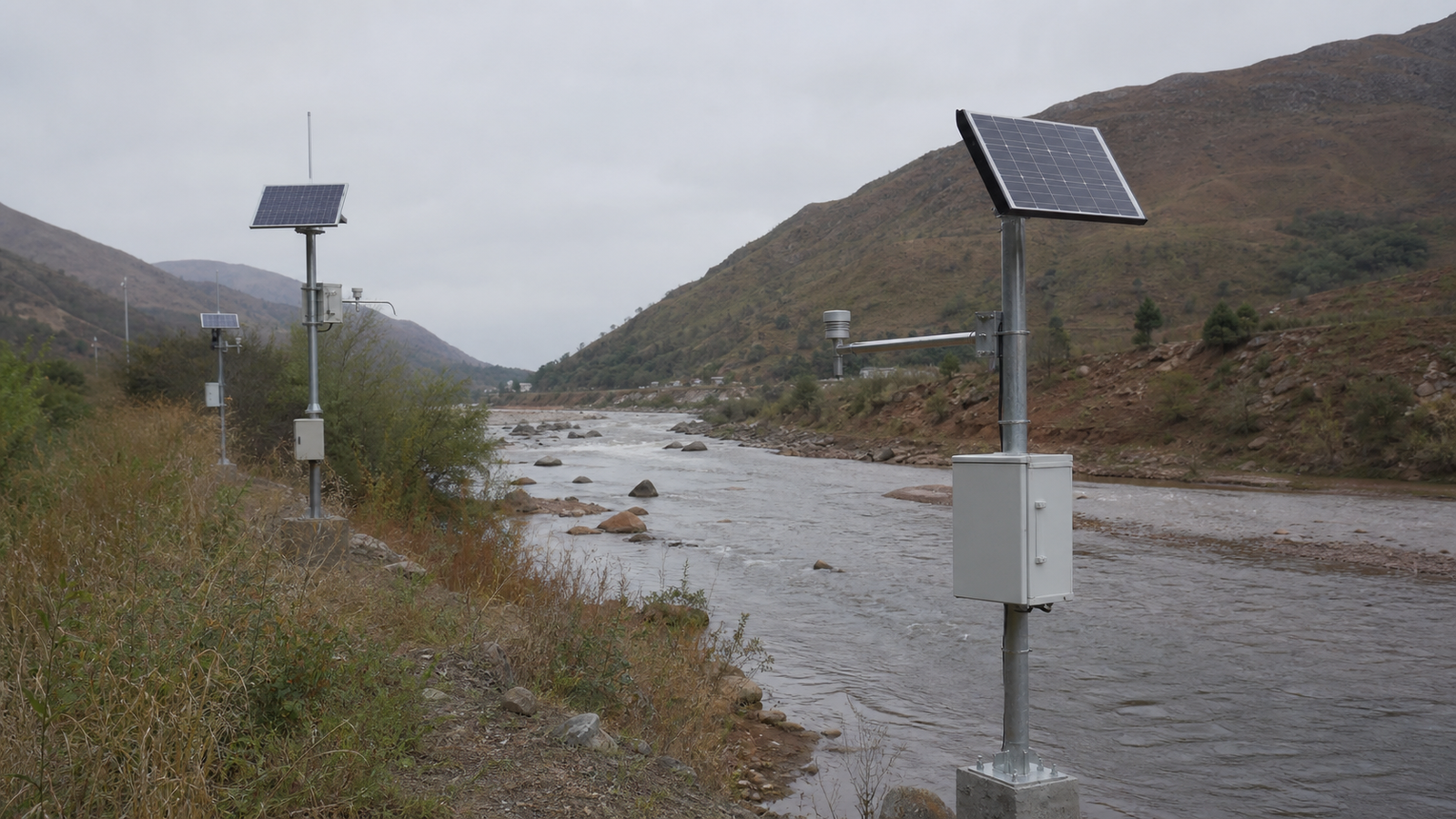 Multiple solar-powered hydrological monitoring stations installed along a wide river valley under an overcast sky.