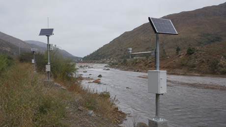 Multiple solar-powered hydrological monitoring stations installed along a wide river valley under an overcast sky.