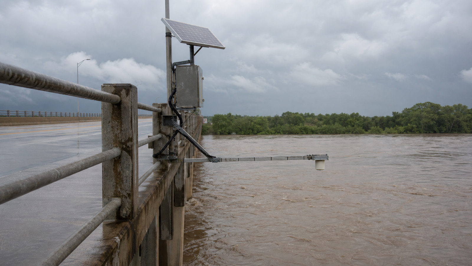A solar-powered radar water level monitoring station mounted on a bridge over a wide river under an overcast sky.