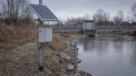 A remote river gauge station with a tilted solar panel on a pole, a weatherproof enclosure and a stage sensor pipe at the water's edge, on an overcast riverbank with a footbridge.