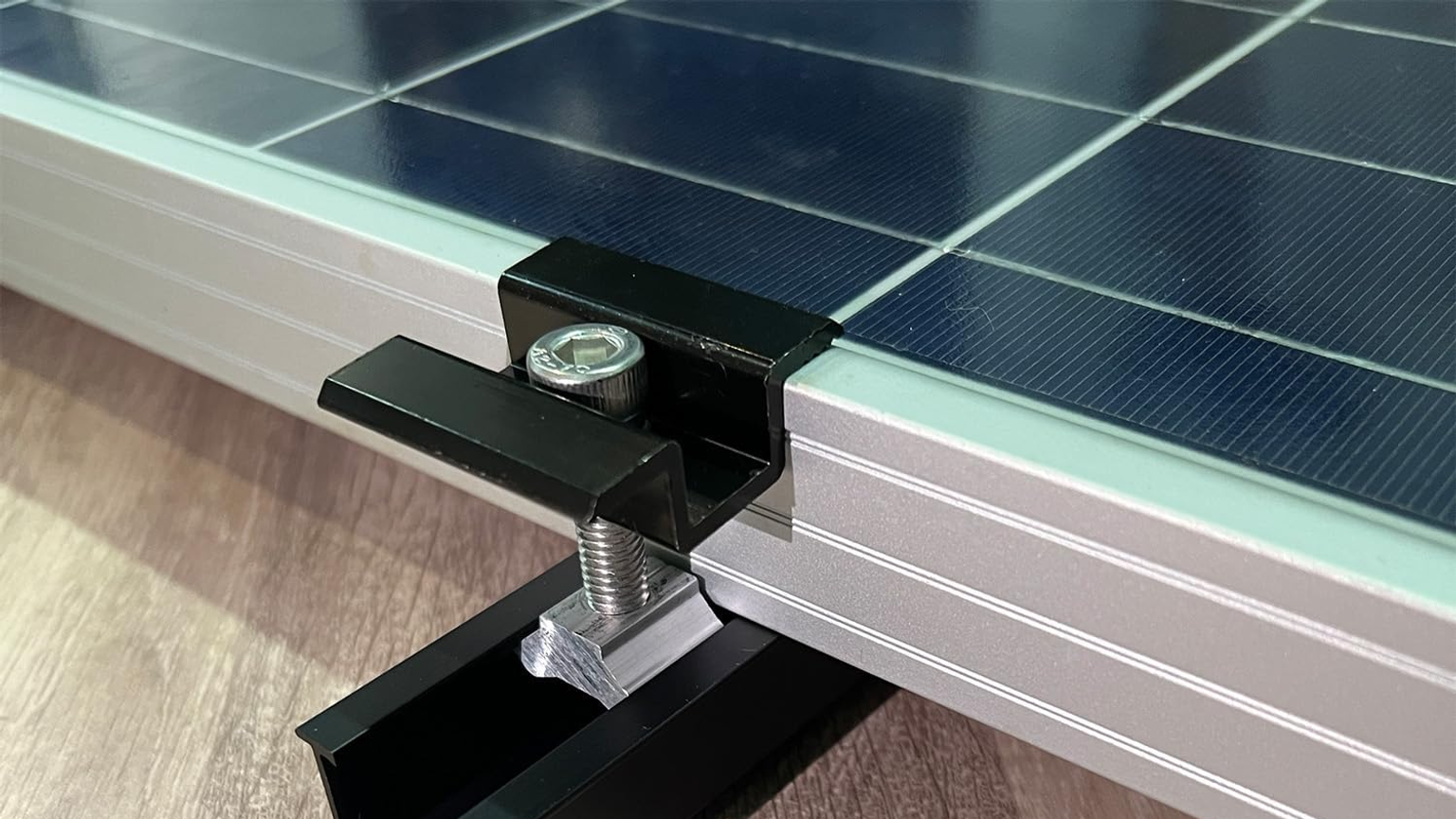 Close-up of a black aluminum short solar rail, sliding nut, end clamp, and stainless fastener securing an unbranded framed panel.