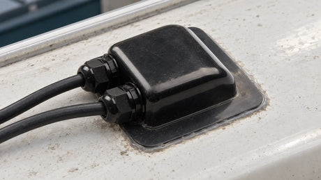 A black double-entry cable housing is mounted on a plain camper roof, with two insulated cables and the surrounding base seal visible.