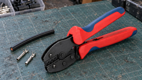 A solar-connector crimping tool with red and blue handles rests beside unpowered cable and contact samples on a workshop mat.