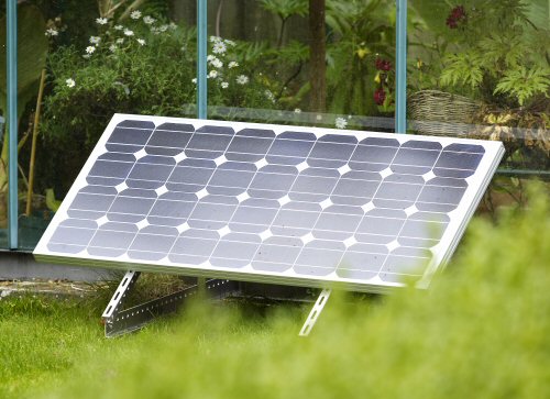 Framed solar panel on a low tilt stand on a garden lawn, set outside a glass greenhouse rather than behind it