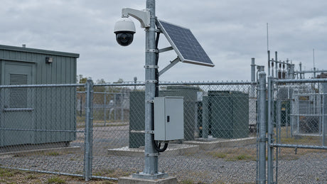 A pole-mounted PTZ camera, solar panel and equipment cabinet form a generic remote monitoring installation at an ordinary fenced utility site.