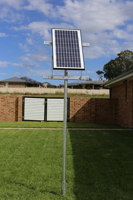 Small rigid solar panel on a galvanized pole in a backyard, the kind of mount used for a shed or coop fan