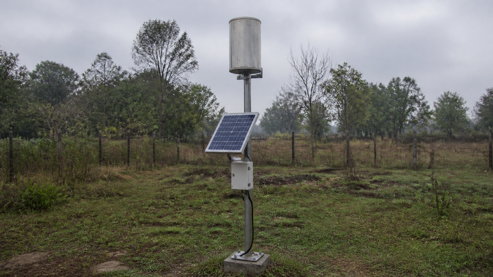A solar-powered rain gauge station with tipping-bucket gauge and small panel mounted on a pole in an open rural clearing under an overcast sky.