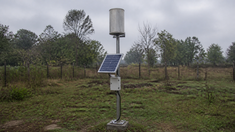 A solar-powered rain gauge station with tipping-bucket gauge and small panel mounted on a pole in an open rural clearing under an overcast sky.