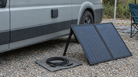 A portable foldable solar panel stands beside an unbranded camper van at an ordinary campsite, with its cable disconnected while the inlet arrangement is reviewed.