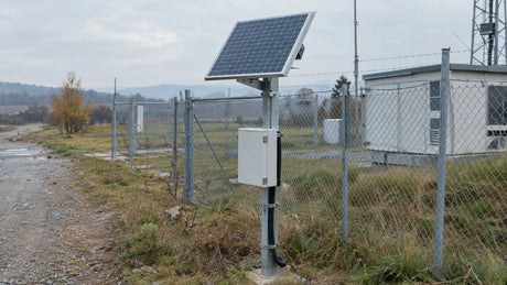 One compact finished PV module securely mounted on a modest post beside an ordinary remote-equipment fence, separate closed equipment enclosure below with protected cable route and fasteners.