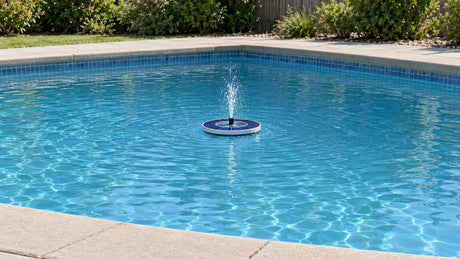 A small floating solar fountain making a low spray in an empty swimming pool, with its top PV surface and the pool edge in view.