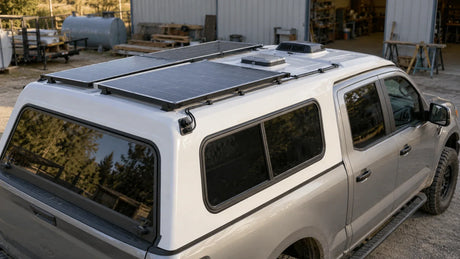 A pickup-mounted camper has a compact rooftop solar array positioned around vents at a plain service yard.