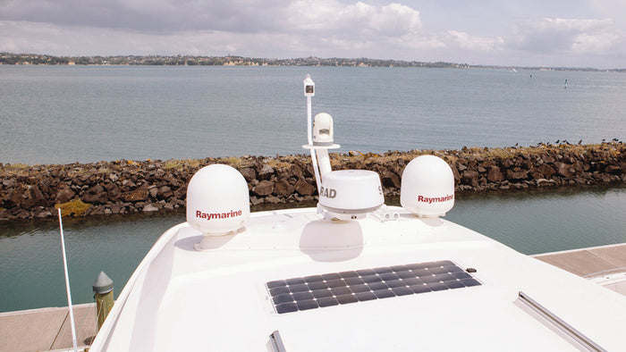A flexible solar panel mounted on the white deck of a boat, with Raymarine radar domes, a RAD antenna, and water in the background.