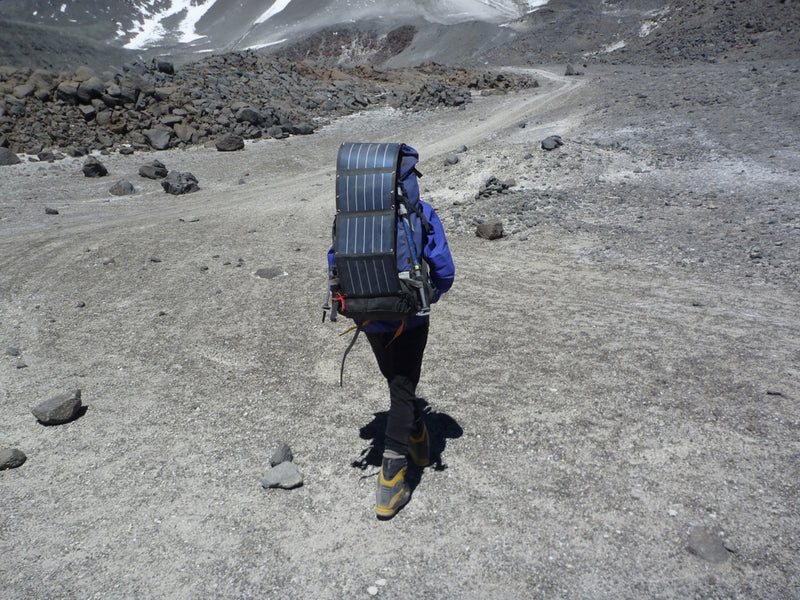 A climber wearing a blue jacket and a large backpack with a solar panel attached, hiking up a barren, rocky mountain trail.