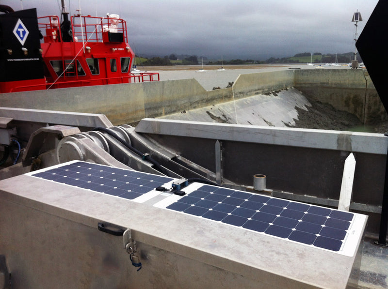 A flexible solar panel installed on a large, gray metal surface on the deck of an industrial boat, with a red structure in the background.
