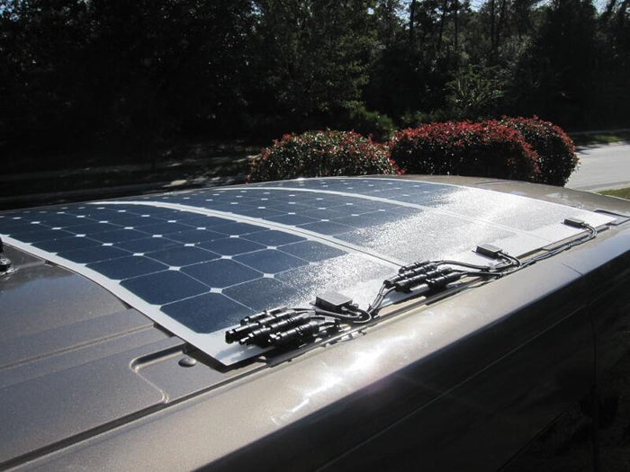 Three flexible solar panels installed on the curved roof of a van, reflecting bright sunlight. The wiring and connectors are visible at the panel edges.