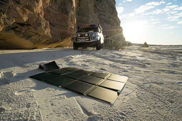A LINKSOLAR folding solar blanket laid out on the sand, charging an off-road vehicle parked in the background by a rocky cliff.