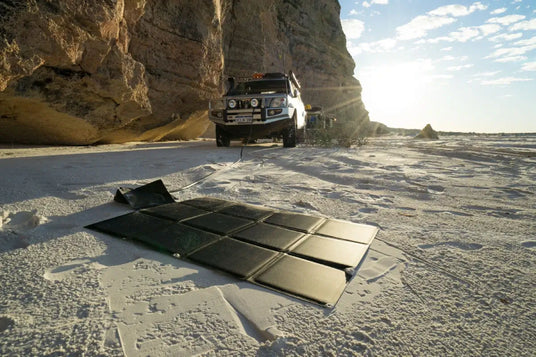 A folding solar blanket laid on white sand, connected to an off-road vehicle parked against a large rock formation in a sunny, arid landscape.