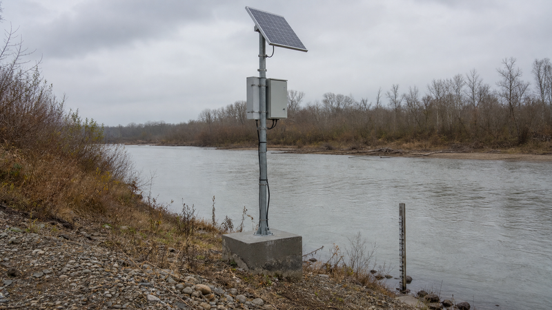 Solar-powered river gauge mounted on a tall pole and concrete foundation above the riverbank, with a staff gauge at the water.