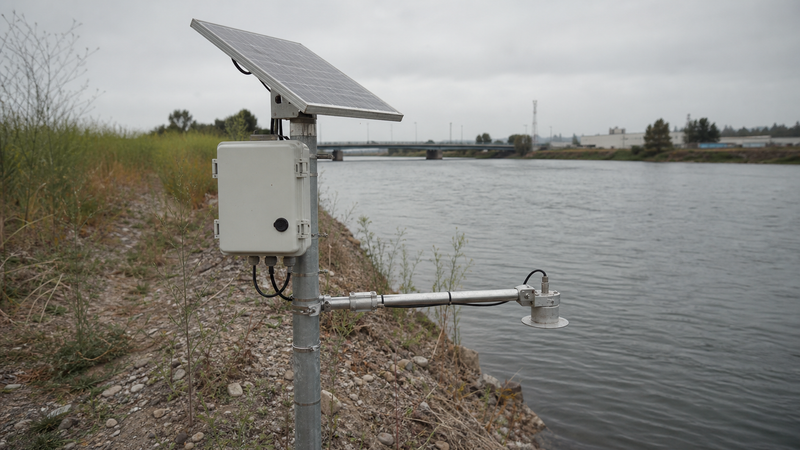A remote water-level monitoring station on a riverbank with a tilted solar panel and weatherproof enclosure on a pole above the flood line and a radar sensor arm over the water.