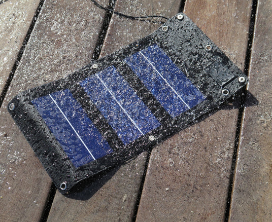 A small, black fabric-edged solar panel covered in water droplets, lying on a wet wooden deck to demonstrate its waterproof quality.